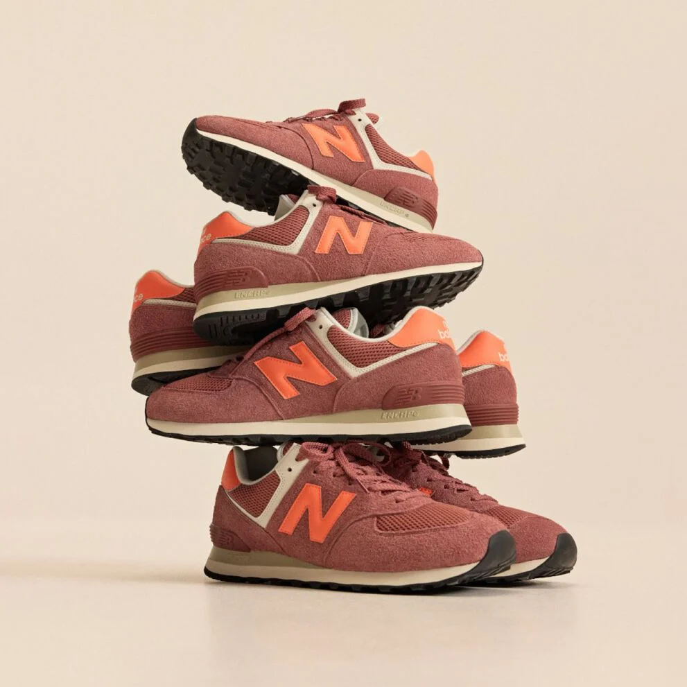 Stack of brick‑red New Balance sneakers with orange accents arranged in a balanced tower against a beige background. 